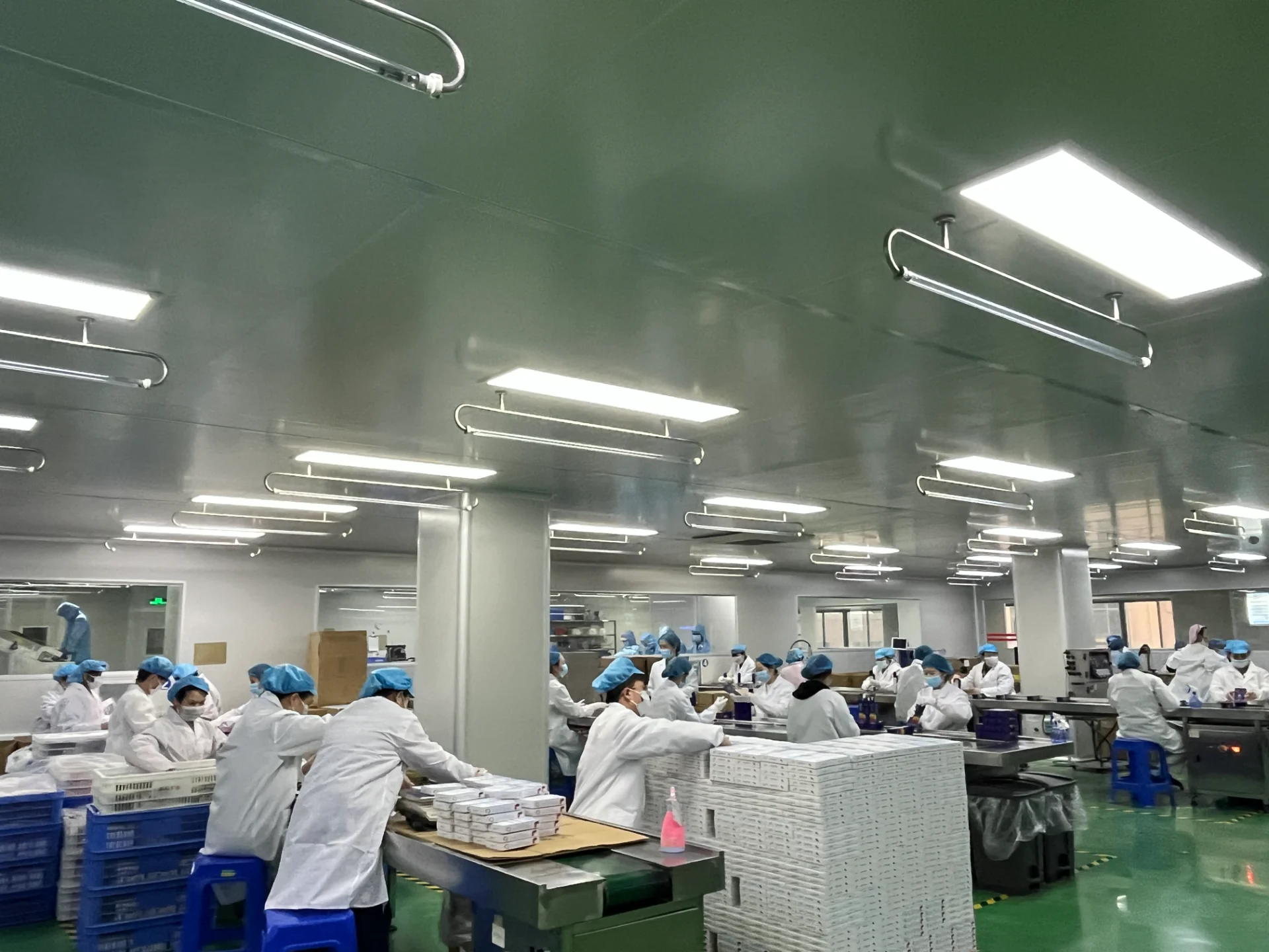Employees in white lab coats and blue hairnets working on an assembly line in a well-lit, clean factory environment.